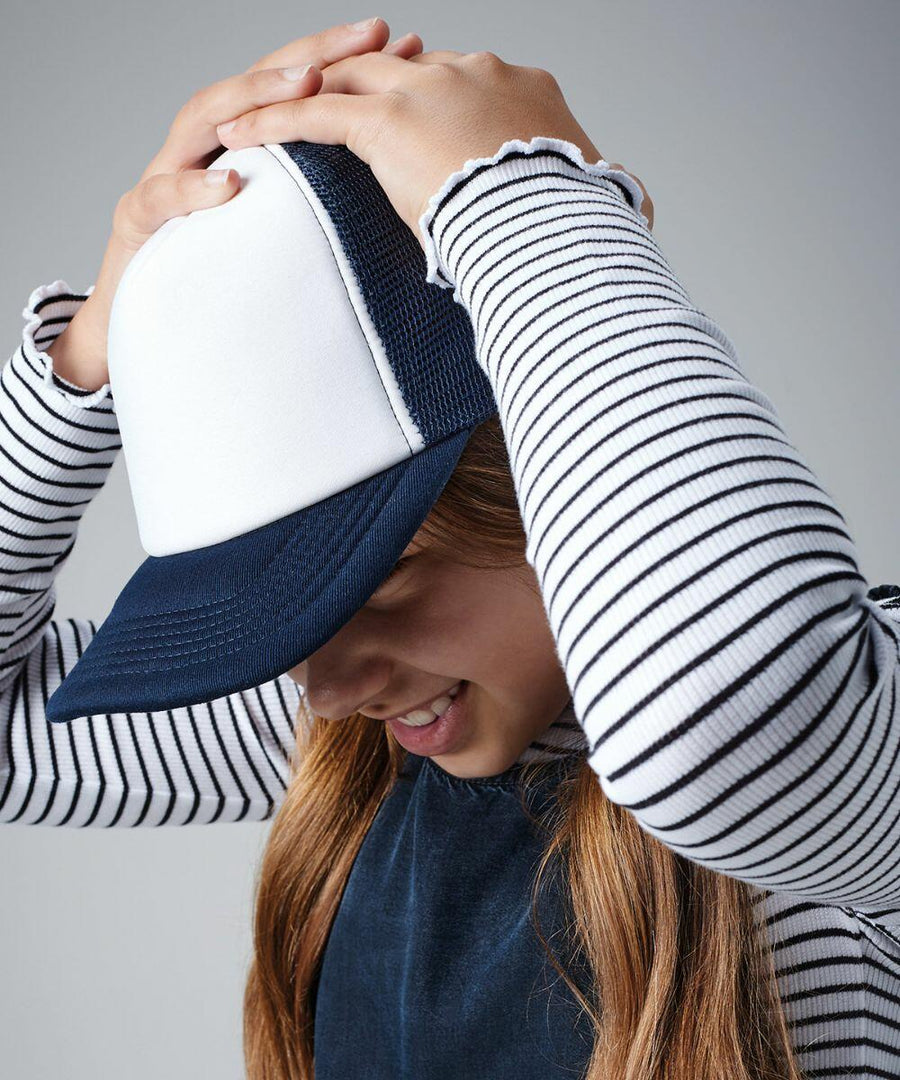 A person wearing a blue and white Beechfield B645B trucker hat and a black apron over a long-sleeved, white and black striped shirt is smiling and holding their hat with both hands. The background is plain and gray.