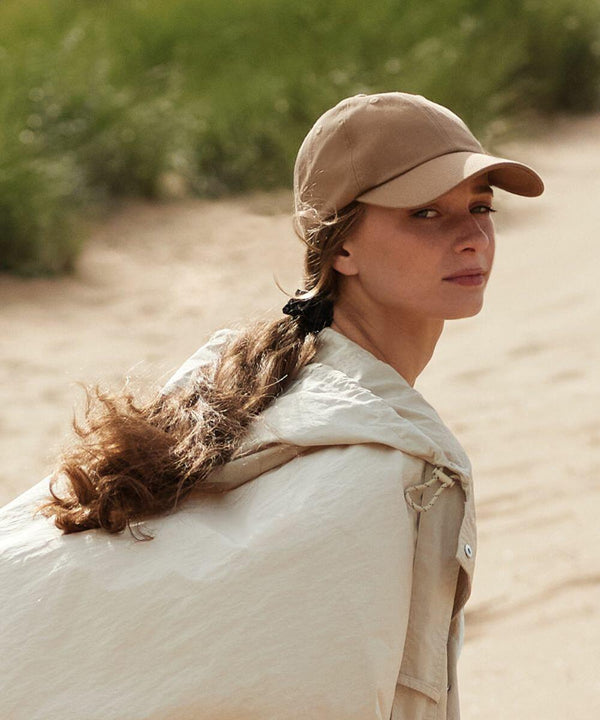 A person with long hair, tied in a ponytail, is wearing a beige Beechfield B652N and a light-colored jacket. They are standing outdoors on a sandy path with green shrubs in the background, looking over their shoulder towards the camera with a neutral expression.