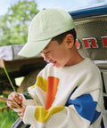 A child in a Beechfield Junior low-profile 6-panel dad cap and a cream jumper with colourful patterns stands by a vehicle grille, holding and examining grass stems.