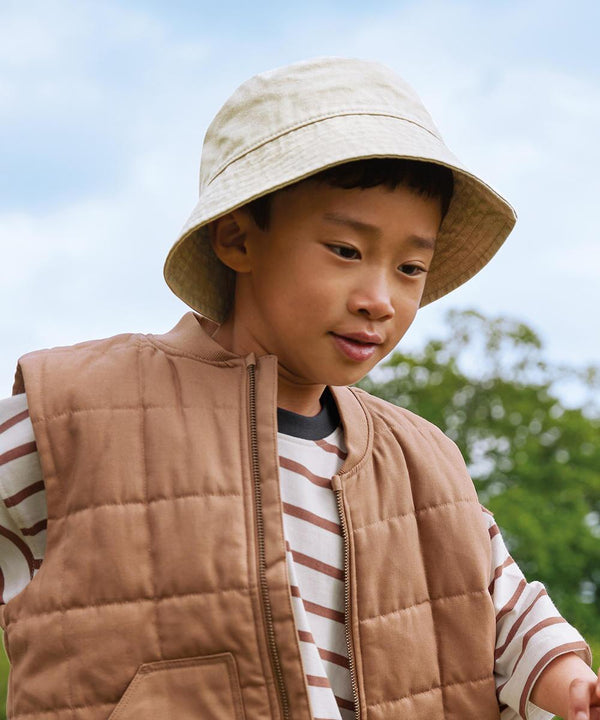 A young child wears the Beechfield Junior Vintage Bucket Hat with a brown quilted gilet and striped shirt, standing outdoors against trees and a blue sky.