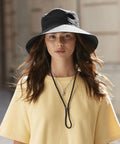 A woman with long brown hair wears a Beechfield wide brim sun hat with a chin strap, a yellow T-shirt, and layered necklaces. She stands outdoors in soft natural light.