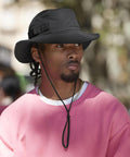 A man wearing the Beechfield Outdoor Adventure Sun Hat with a chin strap and a pink jumper looks to the side in an outdoor, blurred setting.
