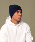 A person wears the Beechfield Organic Cotton Original Cuffed Beanie in navy blue with a white hoodie, looking to the side against a plain, neutral background.