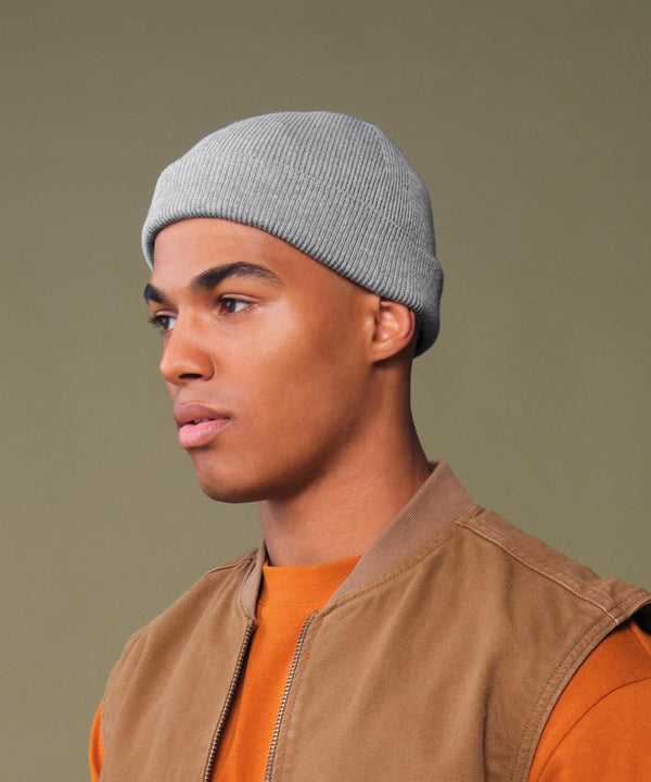 A person in profile wears a Beechfield Organic cotton fisherman beanie in light grey, an orange shirt, and a brown waistcoat against a plain olive green background.