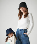 A woman and a child are both wearing Beechfield B90NB bucket hats and casual outfits. The woman is dressed in a white long-sleeve shirt and blue jeans, with one hand in her pocket, while the child is in a white shirt with graphics and blue jeans. They are looking at the camera.
