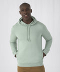 A person wearing a light green B&C Collection BA001 hoodie with the hood down and brown pants, standing with their hands in their pockets. The background is plain white.