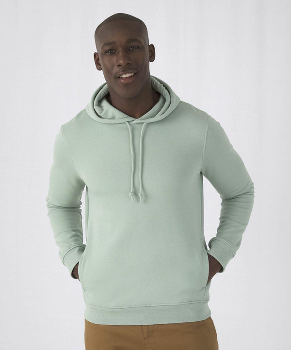 A person wearing a light green B&C Collection BA001 hoodie with the hood down and brown pants, standing with their hands in their pockets. The background is plain white.