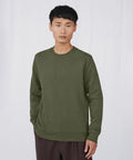 A person with short black hair is wearing an olive green long-sleeve BA010 sweater by B&C Collection and brown pants. They are standing against a plain white background, looking straight ahead with a neutral expression.