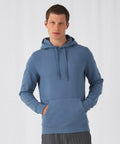 A person with short hair wears a light blue B&C Collection BA011 with a front pocket and drawstrings. They stand against a plain white background, looking directly at the camera, with their hands resting in the hoodie pocket.