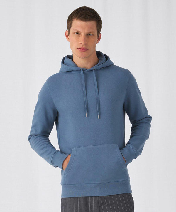 A person with short hair wears a light blue B&C Collection BA011 with a front pocket and drawstrings. They stand against a plain white background, looking directly at the camera, with their hands resting in the hoodie pocket.