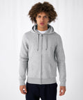 A young person with curly hair is standing against a plain white background, wearing a light gray B&C Collection BA012 zip-up hoodie with drawstrings and front pockets over a white shirt, and dark blue pants. The individual is looking straight at the camera with a neutral expression.