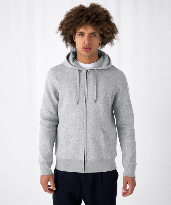 A young person with curly hair is standing against a plain white background, wearing a light gray B&C Collection BA012 zip-up hoodie with drawstrings and front pockets over a white shirt, and dark blue pants. The individual is looking straight at the camera with a neutral expression.