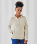 A person with curly hair is wearing a beige B&C Collection BA01F hoodie with a front pocket and blue pants. They are looking at the camera with a neutral expression, standing against a plain white background. One hand is casually in their pocket.