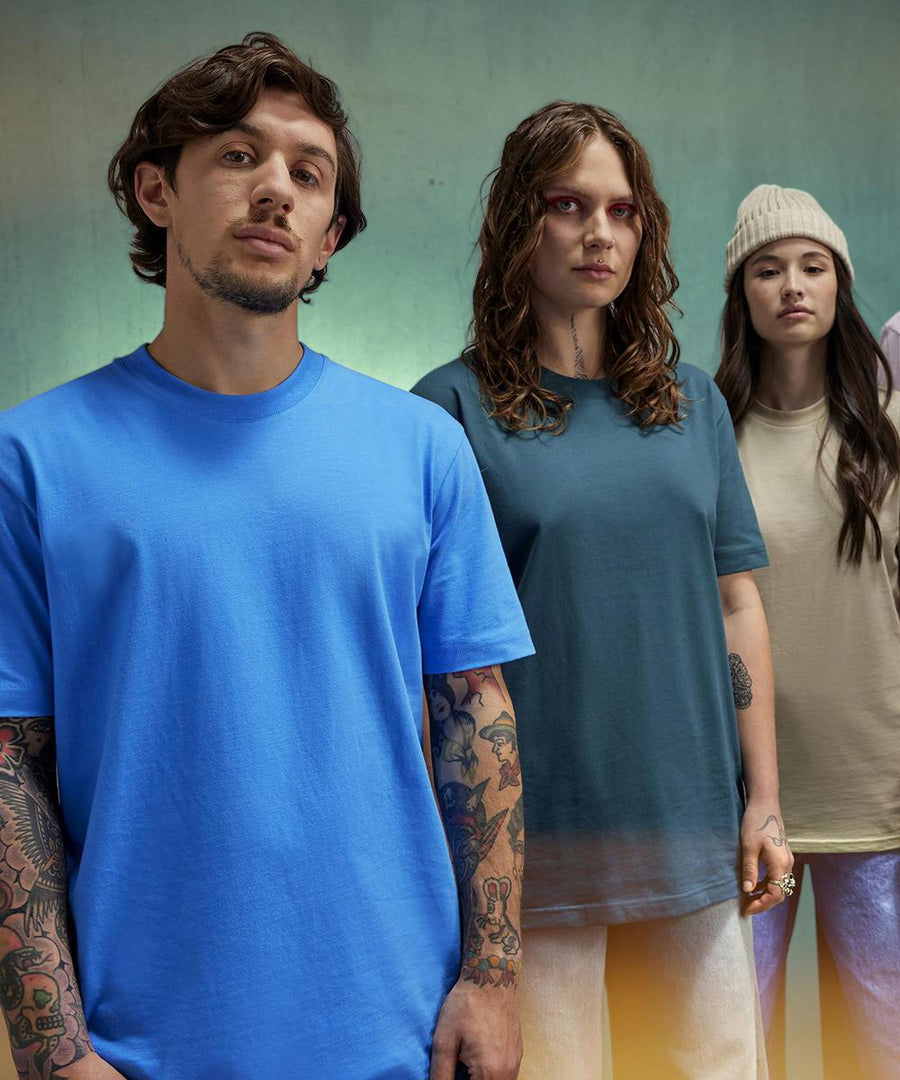 Three people face the camera. The person on the left wears a blue B&C Collection B&C #E220 t-shirt, the centre person has a teal version, and the rightmost wears a beige one with a knitted beanie.