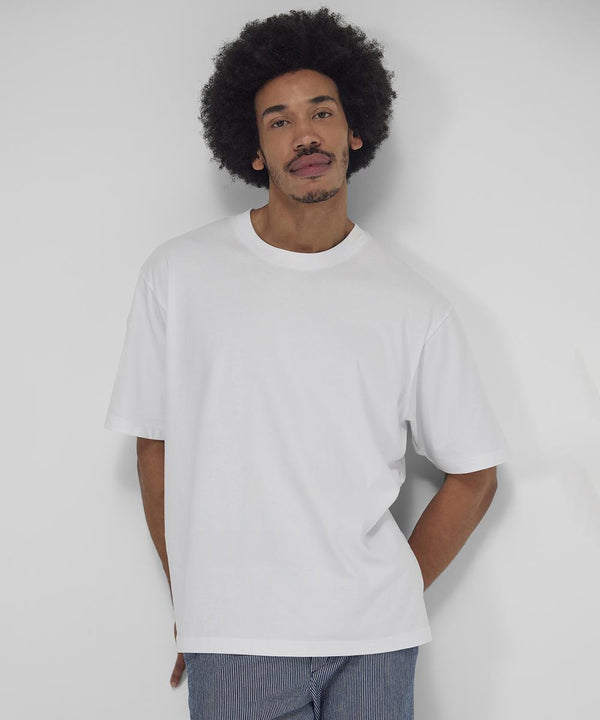 A person with short curly hair stands against a white background, wearing the B&C Collection B&C #E220 Oversized white T-shirt and blue pinstriped trousers, one hand in their pocket, looking at the camera.