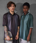 Two people stand by a concrete wall, looking at the camera. One holds a skateboard and has tattoos. Both wear loose B&C Active polo shirts from B&C Collection—one in dark, one in green—with short sleeves and collars.