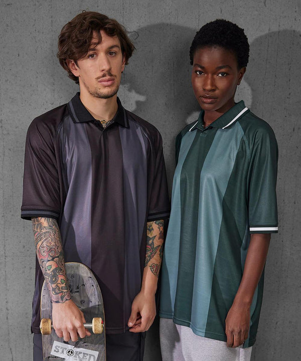 Two people stand by a concrete wall, looking at the camera. One holds a skateboard and has tattoos. Both wear loose B&C Active polo shirts from B&C Collection—one in dark, one in green—with short sleeves and collars.