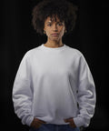 A person with short curly hair wears a B&C Collection Influence crew white sweatshirt and blue jeans, standing against a dark background with hands in their pockets, looking directly at the camera.