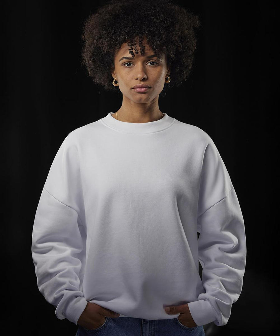 A person with short curly hair wears a B&C Collection Influence crew white sweatshirt and blue jeans, standing against a dark background with hands in their pockets, looking directly at the camera.