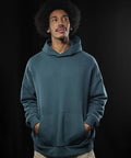 A man wearing the B&C Influence hoodie by B&C Collection stands against a dark background with his hands in the front pocket, looking slightly upwards and to the side.