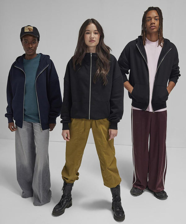 Three people face forwards against a plain light background, each wearing the B&C Collection B&C Influence zip hoody. One wears a cap, another boots, and the third has plaits; all pair their hoodies with loose trousers and casual shoes.