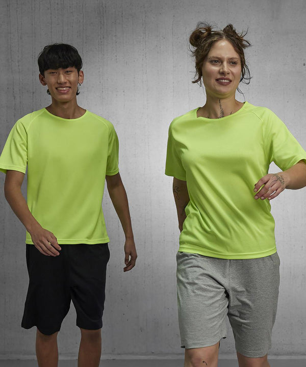 Two people in B&C Collection’s B&C #Active T bright yellow short-sleeve shirts and athletic shorts walk indoors against a plain grey concrete wall, appearing in motion and looking ahead.