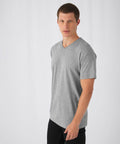A man with short brown hair and fair skin is standing against a plain white background. He is wearing a light grey B&C Collection BA108 V-neck T-shirt and black pants, looking slightly to the side while facing the camera.