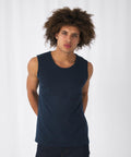 A person with curly hair stands facing forward against a plain white background. They are wearing a dark blue sleeveless B&C Collection BA110 shirt and dark pants, with their arms relaxed behind their back. Their expression is neutral.
