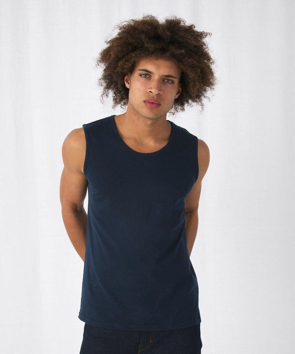 A person with curly hair stands facing forward against a plain white background. They are wearing a dark blue sleeveless B&C Collection BA110 shirt and dark pants, with their arms relaxed behind their back. Their expression is neutral.