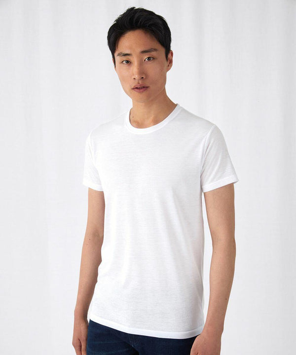 A person with short black hair is standing in front of a plain white background, wearing a B&C Collection BA123 plain white t-shirt and dark pants. They have a neutral expression and are looking directly at the camera.
