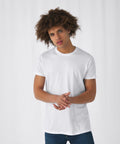 A person with curly hair stands against a plain, light-colored background. They wear a plain white B&C Collection BA210 T-shirt and blue jeans, with their hands clasped together in front of them. The expression on their face appears neutral.