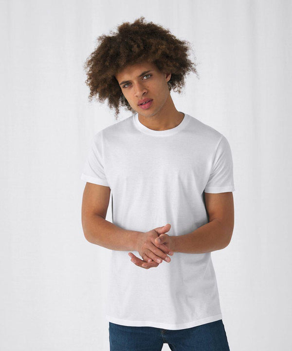 A person with curly hair stands against a plain, light-colored background. They wear a plain white B&C Collection BA210 T-shirt and blue jeans, with their hands clasped together in front of them. The expression on their face appears neutral.