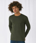 A person with curly hair is standing against a plain white background. They are wearing a long-sleeve, dark green B&C Collection BA211 shirt and blue jeans. Their hands are tucked behind their back, and they are looking slightly to the side with a calm expression.