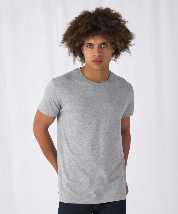 A person with curly hair is standing against a plain white backdrop. They are wearing a plain gray BA212 T-shirt by B&C Collection and dark pants. The individual is looking directly into the camera with a neutral expression.