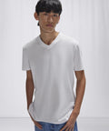 A person with straight dark hair wears the B&C Collection B&C #E150 V plain white V-neck T-shirt and blue jeans, standing against a light background with hands in their pockets.