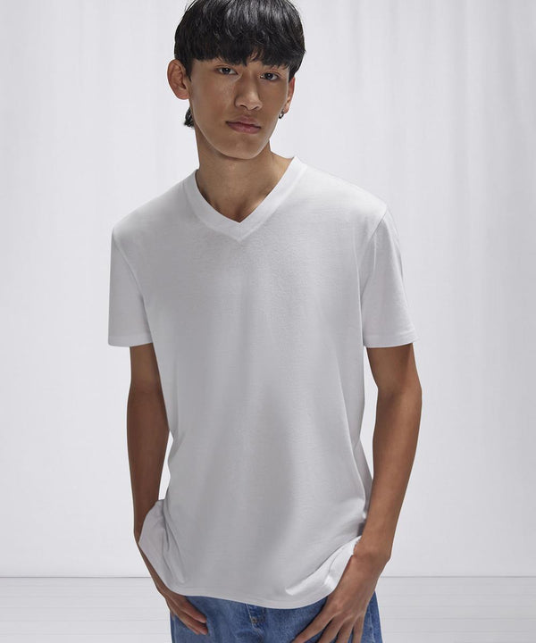 A person with straight dark hair wears the B&C Collection B&C #E150 V plain white V-neck T-shirt and blue jeans, standing against a light background with hands in their pockets.