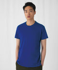 A person with short black hair is standing against a plain white background. They are wearing a plain blue BA220 T-shirt from B&C Collection and khaki pants. The individual's hands are in their pockets, and they have a neutral expression on their face.