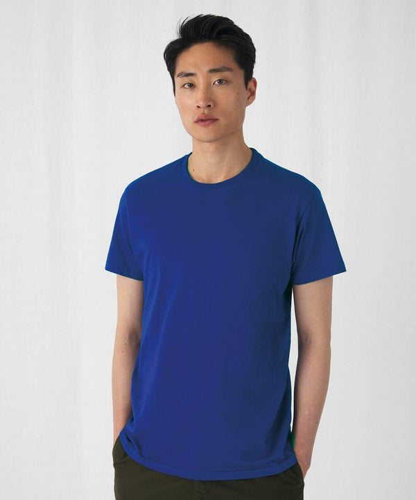 A person with short black hair is standing against a plain white background. They are wearing a plain blue BA220 T-shirt from B&C Collection and khaki pants. The individual's hands are in their pockets, and they have a neutral expression on their face.
