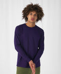A person with curly hair is standing against a white background, wearing a plain, long-sleeve purple BA221 shirt from B&C Collection and olive green pants. Their hands are clasped together in front of them, and they have a neutral expression on their face.