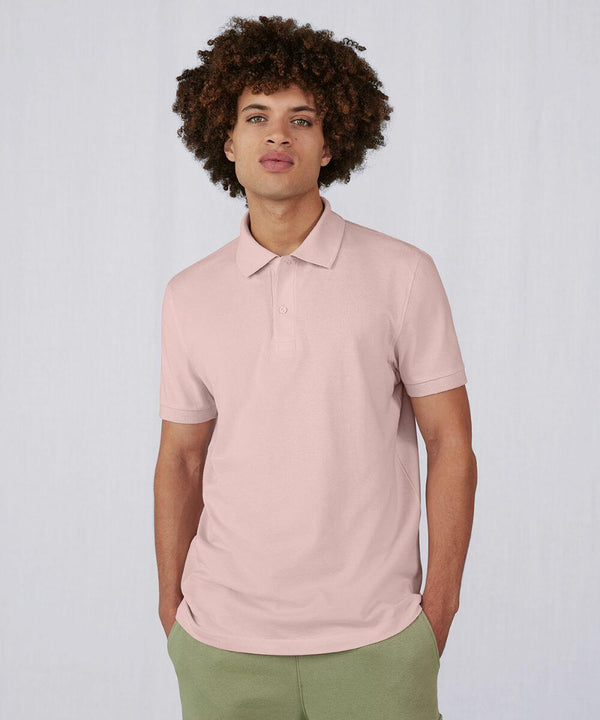 A young man with curly hair stands against a plain, light-colored background. He is wearing a pale pink BA261 polo shirt from B&C Collection and green pants. His hands are casually in his pockets, and he is looking directly at the camera with a neutral expression.