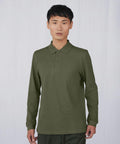 A person with short black hair is wearing a long-sleeve, olive-green BA262 polo shirt by B&C Collection. They are standing against a plain, light gray background, looking directly at the camera with a neutral expression.