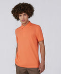 A person with curly hair is wearing an orange B&C Collection BA263 polo shirt and brown pants. They stand against a plain, light gray background, looking forward with their left hand in their pocket.