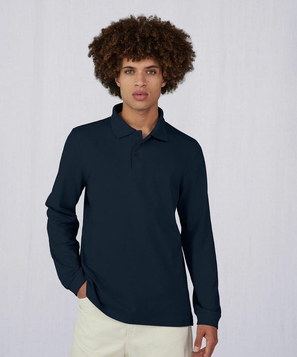 A person with curly hair is wearing a dark blue long-sleeved B&C Collection BA264 polo shirt and light-colored pants. Their left hand is in their pocket, and they are standing against a plain, light-colored background.