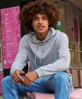 Someone with curly hair wears a B&C #Hoodie by B&C Collection in light grey and blue jeans, sitting on a pink chair outside a shop with pink menus displayed on the wall behind them.