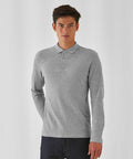 A person with short dark hair is standing against a plain white background, wearing a light gray long-sleeved BA290 polo shirt with a collar and three buttons from B&C Collection, paired with dark jeans. Their arms are relaxed by their sides, and they are looking directly at the camera.