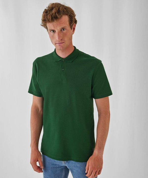 A person with short, curly hair wearing a green BA306 polo shirt from B&C Collection and blue jeans stands in front of a plain white background, looking at the camera with a neutral expression.