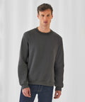 A person stands against a plain white background, wearing a dark gray, long-sleeved B&C Collection BA404 sweatshirt and blue jeans. They have short hair and are looking directly at the camera with a neutral expression.