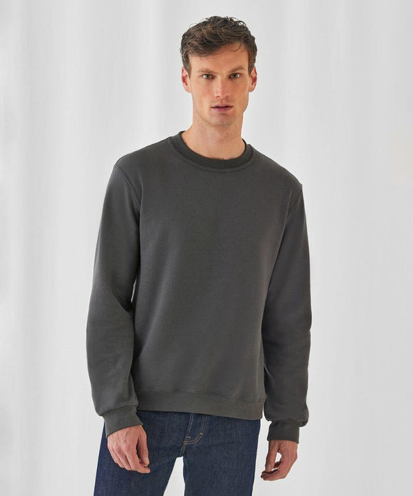 A person stands against a plain white background, wearing a dark gray, long-sleeved B&C Collection BA404 sweatshirt and blue jeans. They have short hair and are looking directly at the camera with a neutral expression.