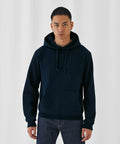 A young person stands against a plain white background wearing a dark navy blue B&C Collection BA405 with the hood down and hands in pockets of blue jeans. The hoodie has drawstrings at the neck and a front kangaroo pocket. They have a neutral expression and earrings in both ears.