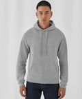 A person with short hair is wearing a B&C Collection BA411 light gray hoodie with a front pocket and dark jeans. They are standing against a plain white background, looking directly at the camera with a neutral expression.